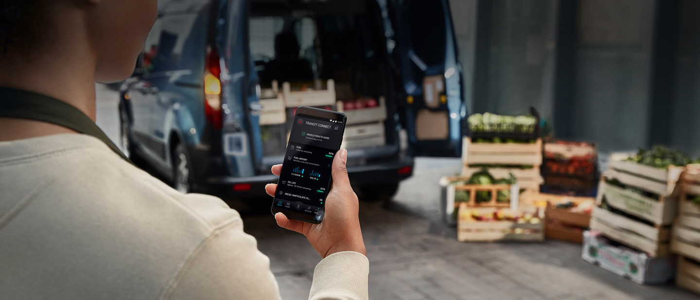 Person kigger på sin telefon med Ford app. I baggrunden holder en Ford Transit Connect læsses med fødevarer parkeret.