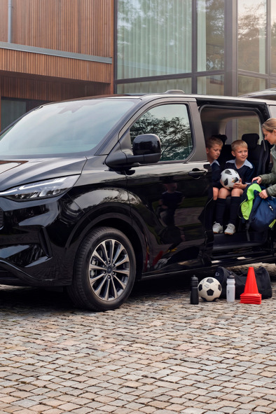 En kvinde hjælper nogle drenge i fodboldtrøjer, mens de stiger ud af en sort varevogn, der holder parkeret på en brostensbelagt indkørsel, med sportsudstyr lige ved siden af.