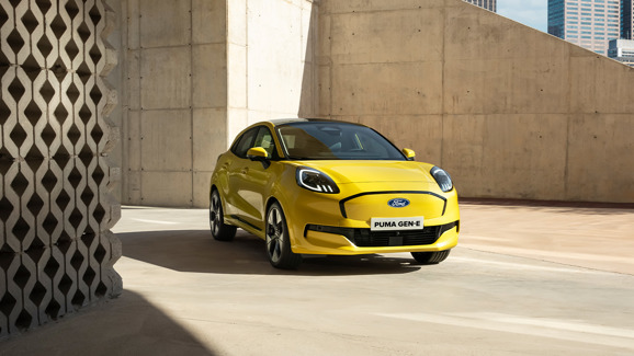 En gul Ford Puma Gen-E er parkeret udenfor ved betonvægge med geometriske mønstre, mod en urban baggrund og et glimt af byens skyline.
