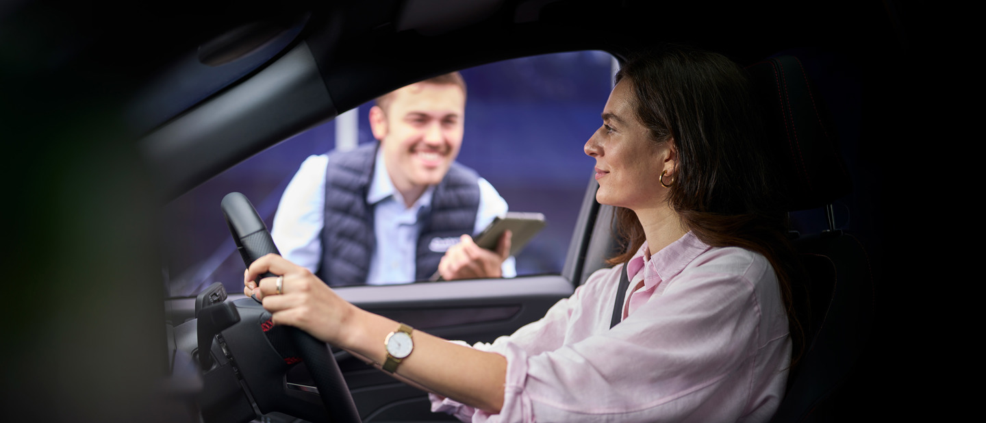 En kvinde i en bil taler med en smilende Ford Randers Servicepartner medarbejder, der står uden for bilvinduet med en tablet i hånden.
