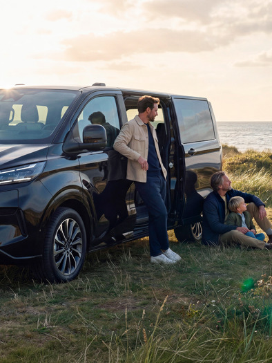 En gruppe mennesker står og sidder ved siden af en parkeret Ford E-Tourneo Custom-varevogn i et græsklædt kystområde under en delvis overskyet himmel.