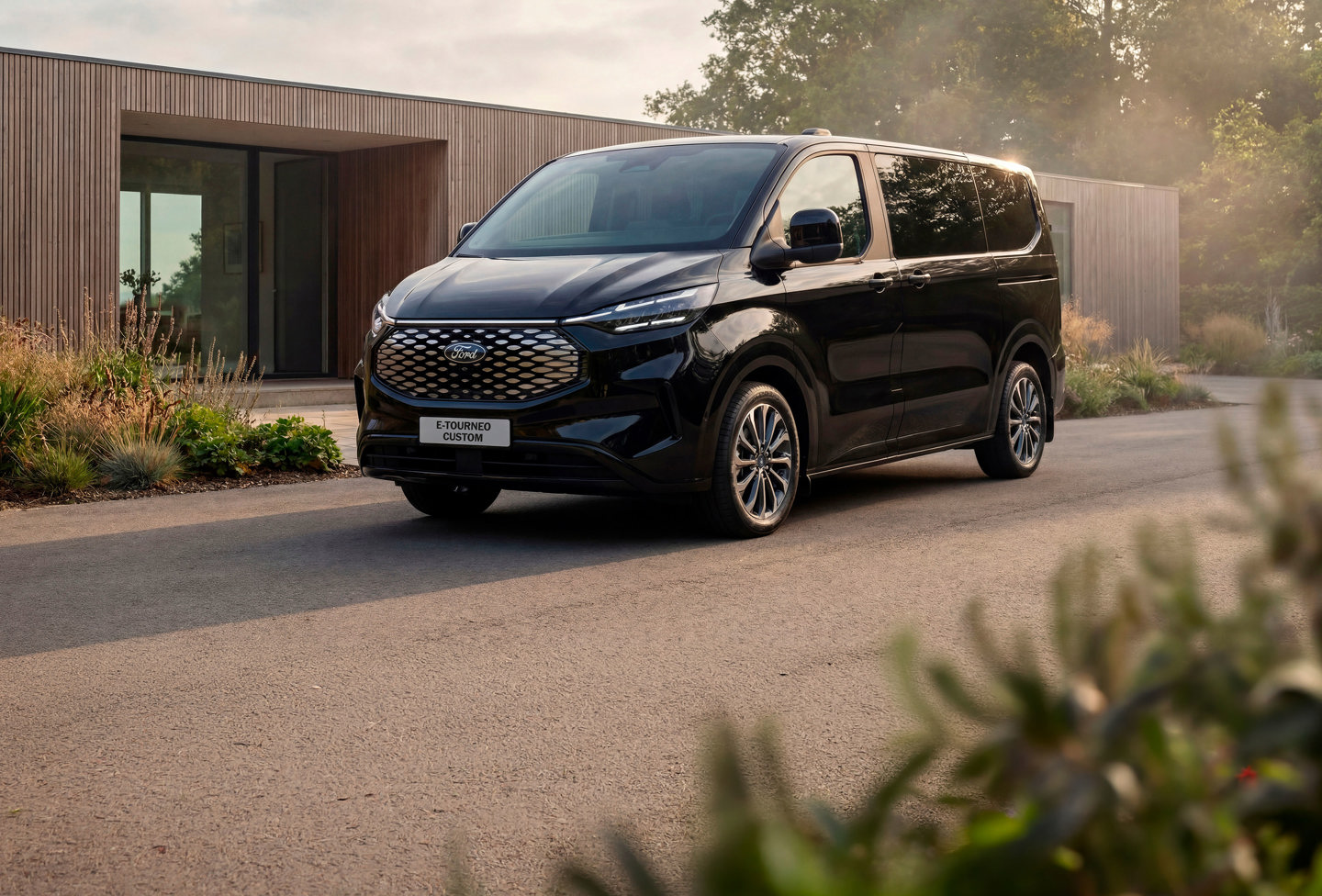 En sort Ford E-Tourneo Custom-varevogn, der holdt parkeret i indkørslen foran et moderne hus med velplejet haveanlæg.