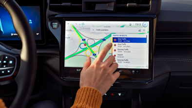 En person bruger bilens navigationssystem med touchscreen, der viser et kort med flere rutevalg og trafikadvarsler.