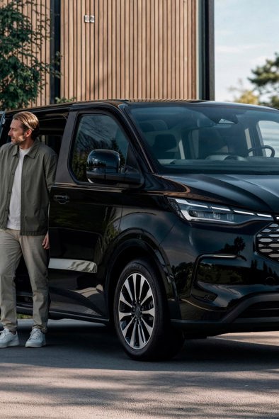 To personer står ved siden af en sort Ford E-Tourneo Custom, der holder parkeret udenfor, og den ene af dem trækker en kuffert efter sig.