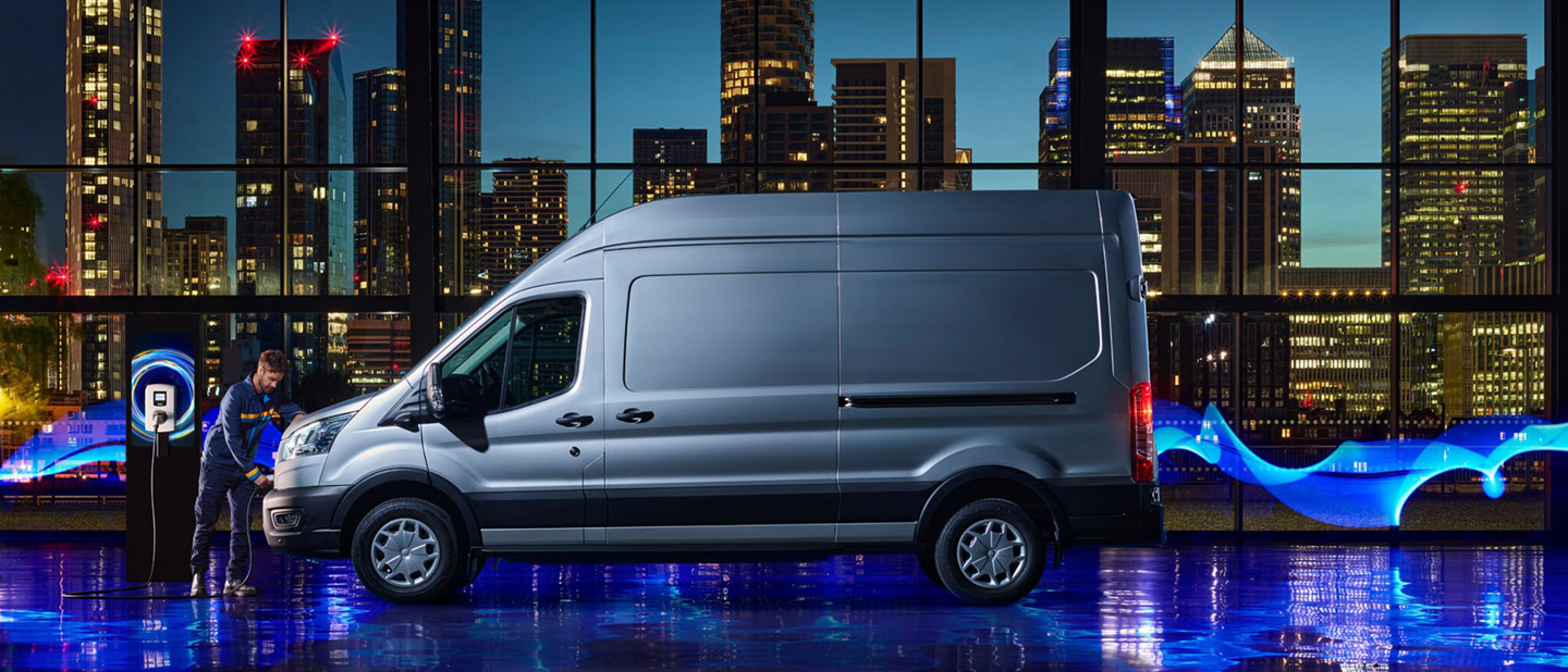 Ford E-Transit set fra siden i udstillingslokale med ambient lys og en mand med et ladekabel.