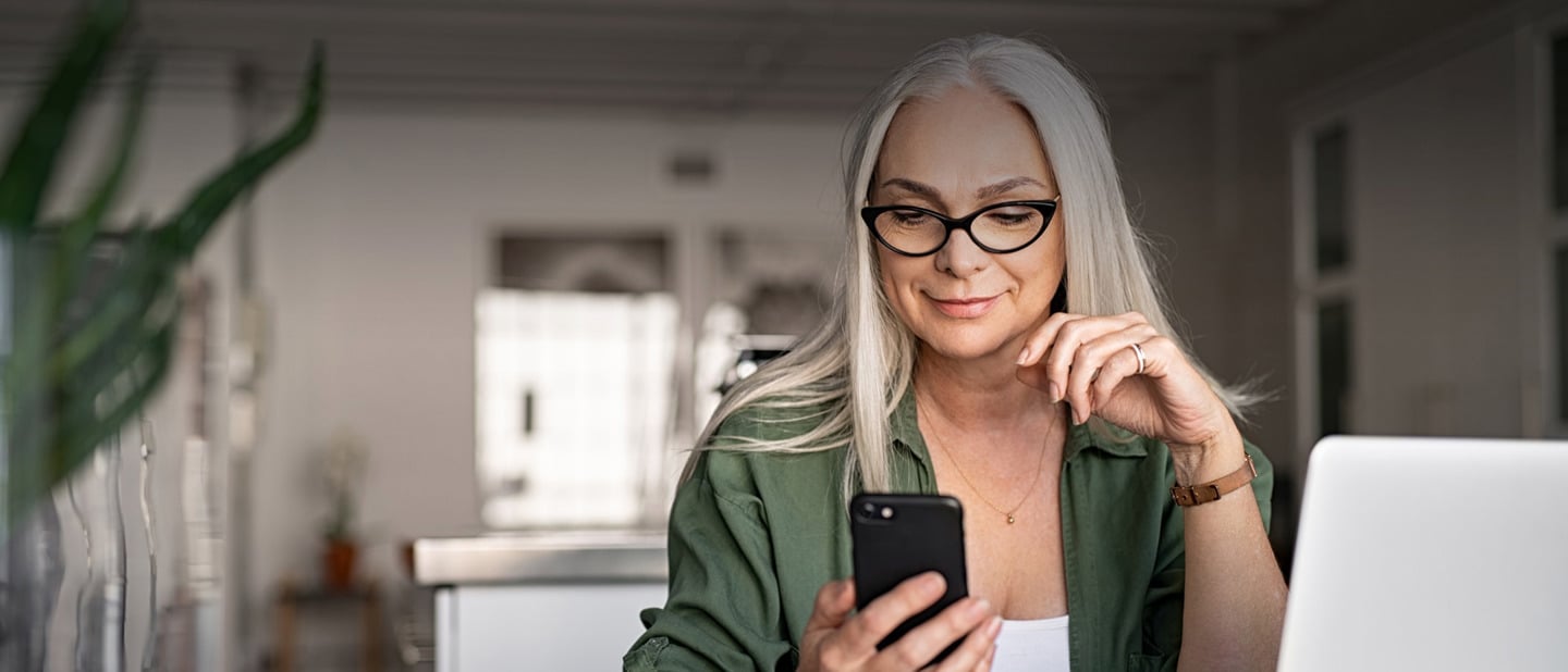 Kvinde med briller sidder ved computer i sit hjem og kigger på sin telefon