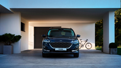 En blå Ford Kuga SUV er parkeret under en moderne carport med en cykel lænet op ad væggen i baggrunden.
