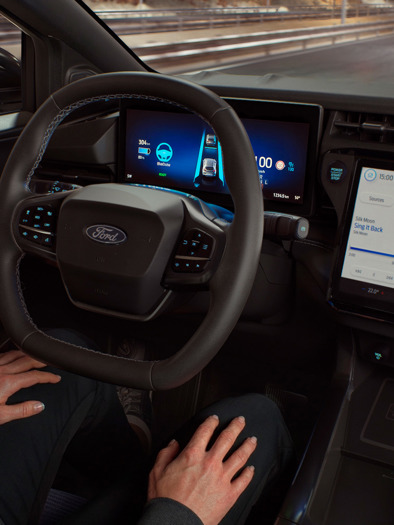 Man sidder trygt i førersædet i en Ford, som kører af sig selv; skærme på instrumentbrættet viser tydeligt navigation og musik og fremhæver avanceret teknologi.