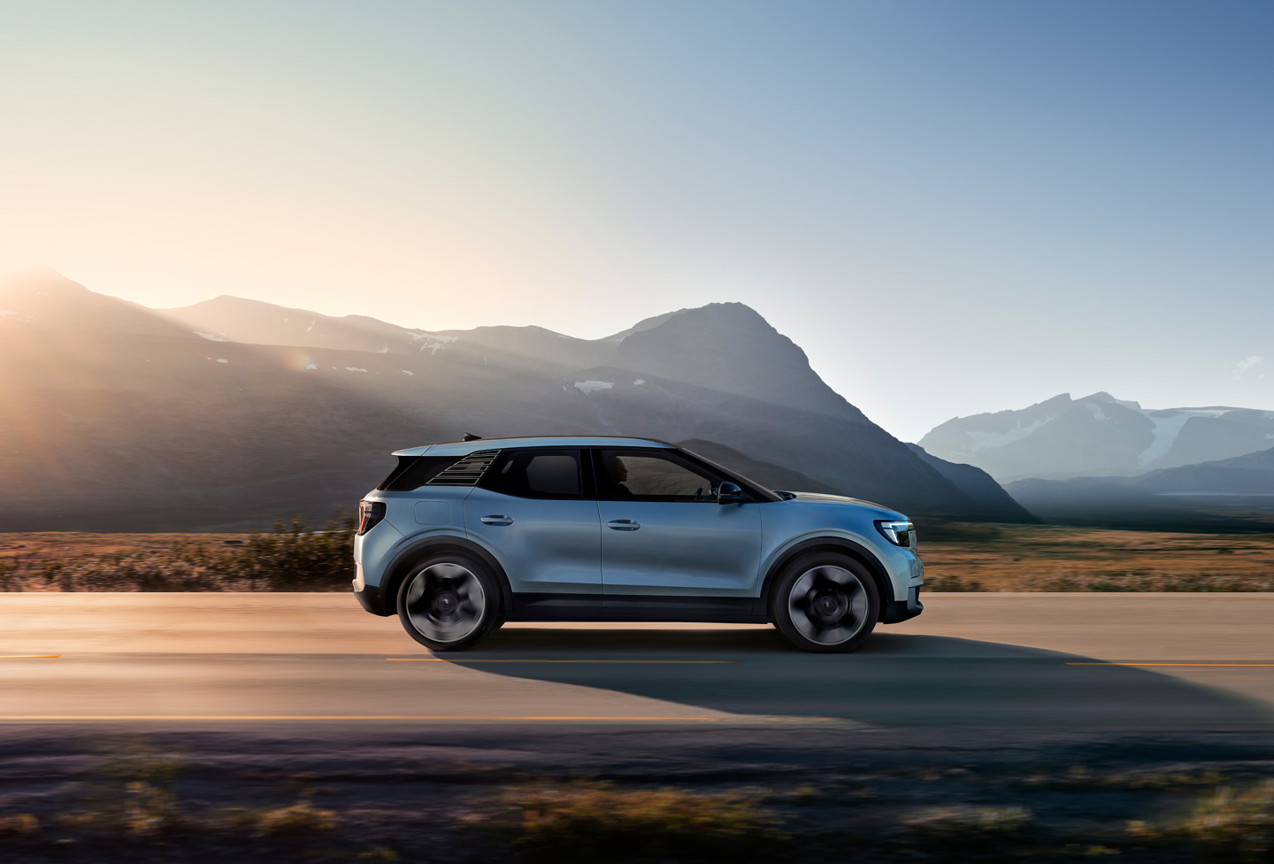 En sølvfarvet SUV kører ad en vej med bjerge i baggrunden og solen, der står op bag dem.