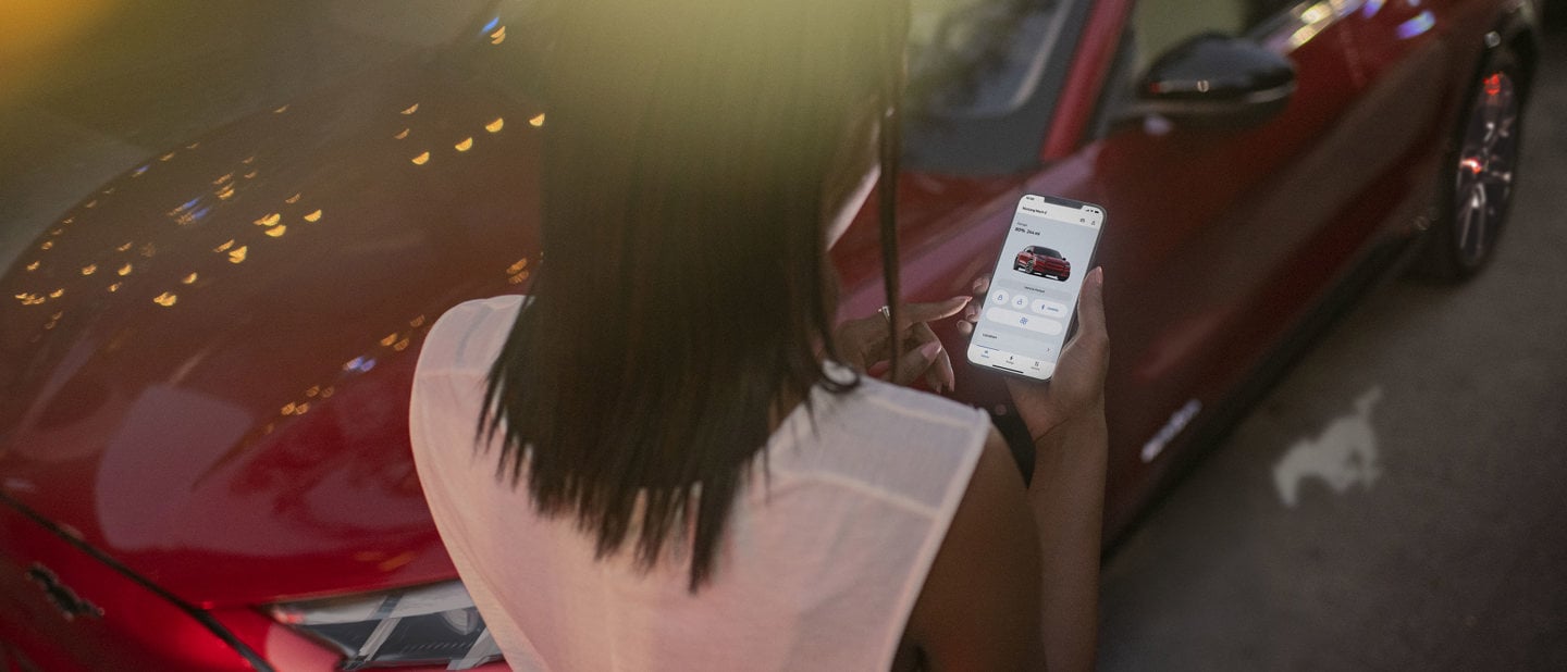 Person står ved rød Ford Mach-E EV med smartphone med FordPass appen åben