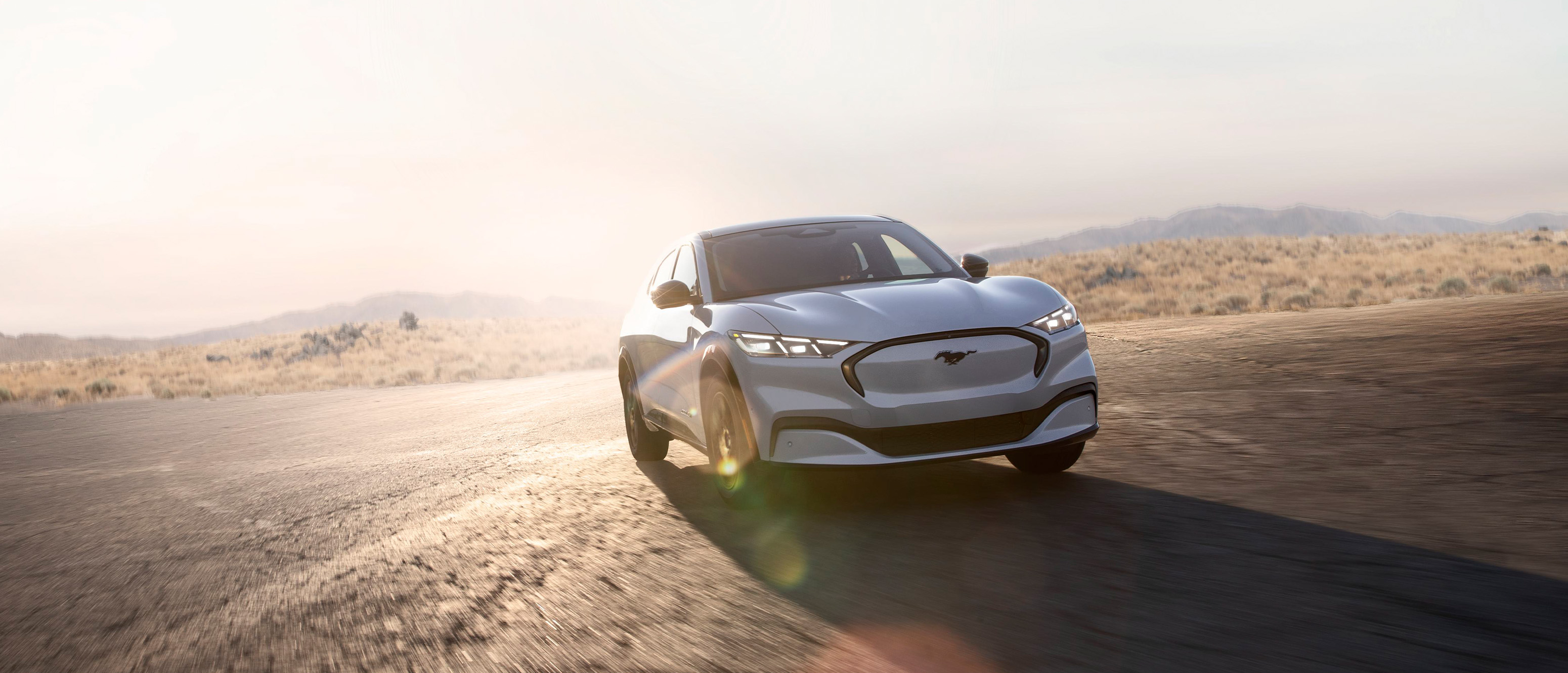 En hvid Ford Mustang Mach-E elektrisk SUV kører på en tom vej med et solbeskinnet landskab i baggrunden.