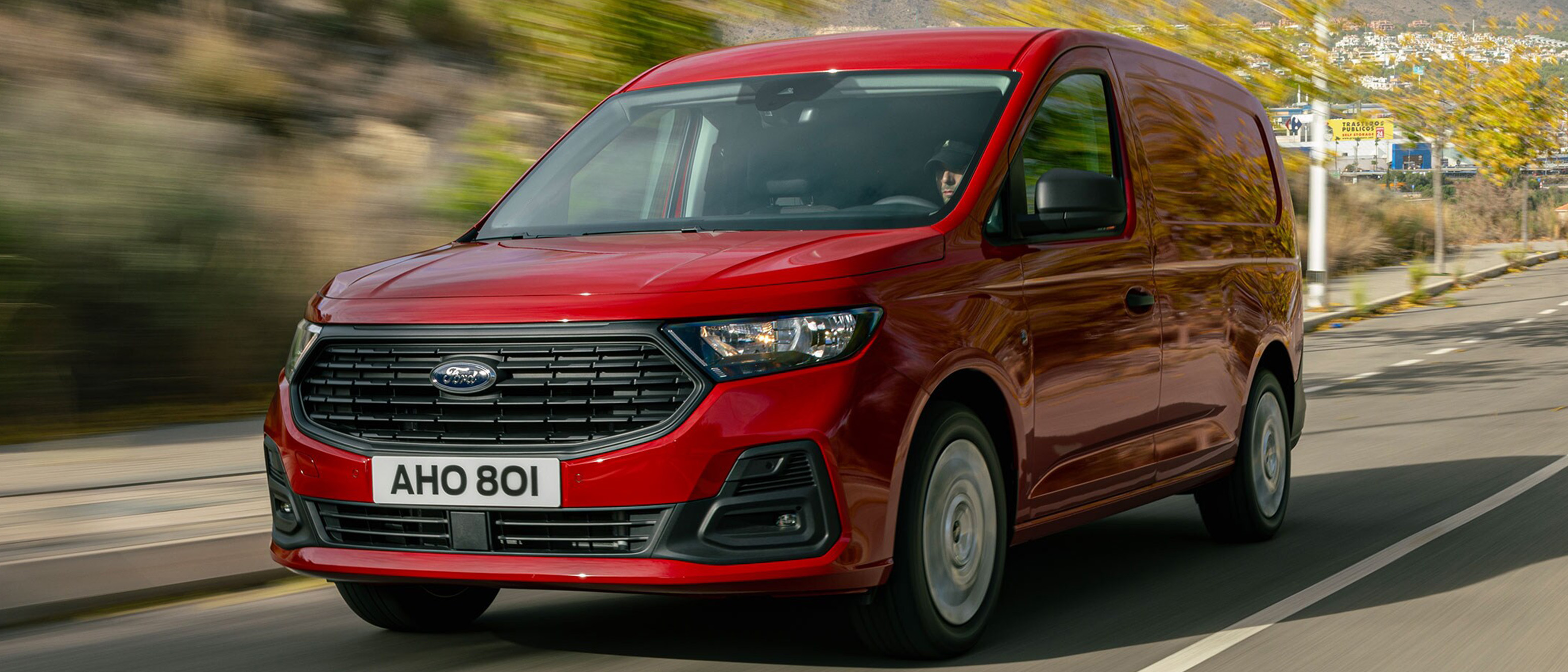 Rød Ford Transit Connect kørende på vej set skråt forfra