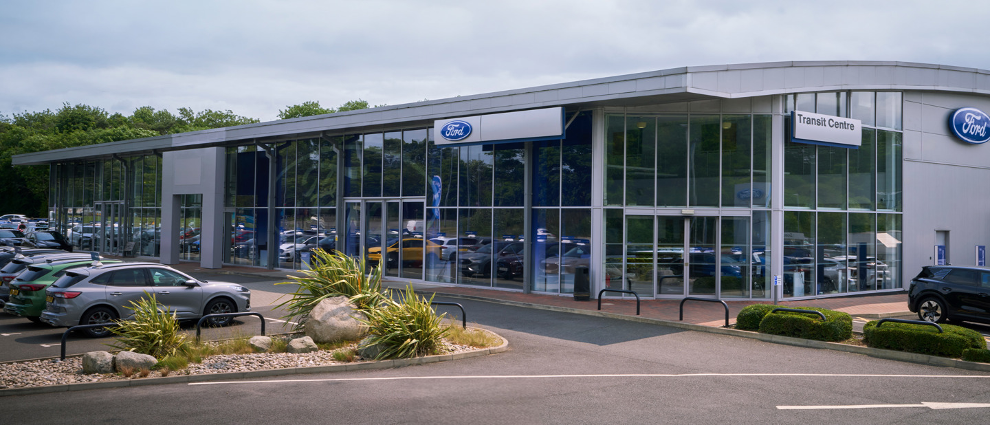 Moderne Ford Risskov bilforhandler med store glasvinduer, flere parkerede biler udenfor og grønne områder omkring bygningen.