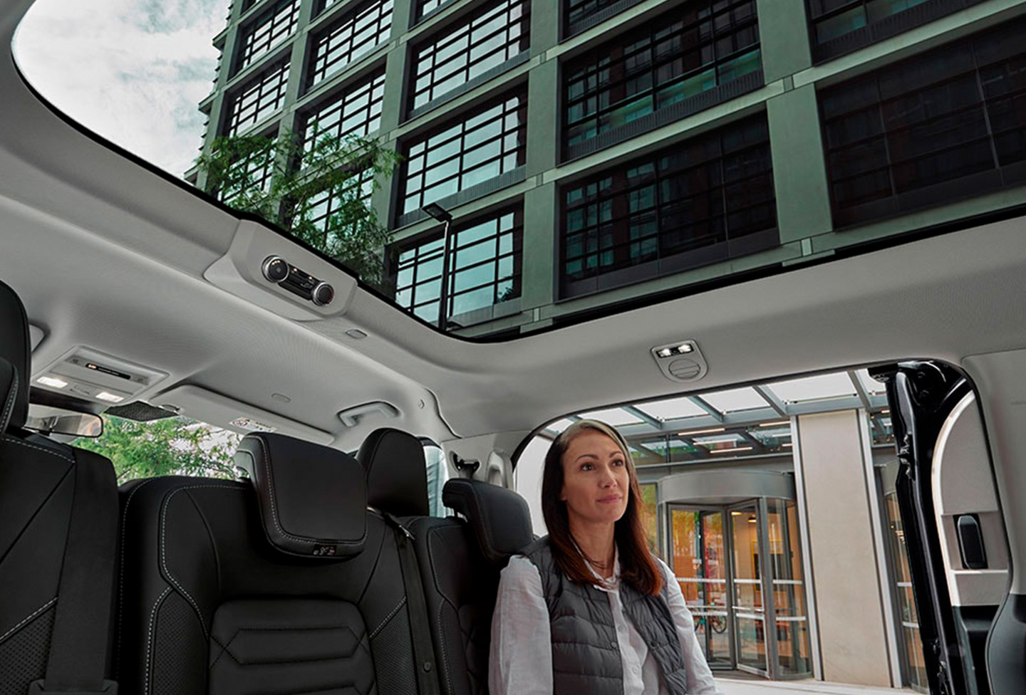 En kvinde sidder på bagsædet i en bil med panoramatag, der holder parkeret tæt på en moderne bygning af glas og stål.