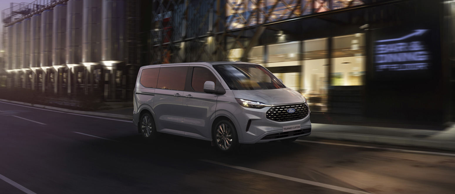 En sølvfarvet Ford Tourneo Custom varebil kører på en gade i byen i skumringen og passerer moderne bygninger med glasfacader.