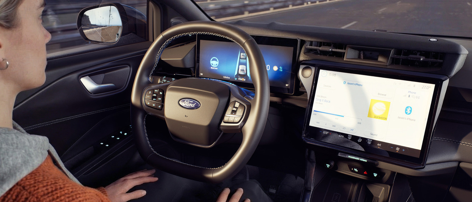 En person sidder i førersædet i en Ford med hænderne væk fra rattet; man ser et digitalt instrumentbræt og en stor touchscreen i midten.
