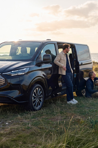 En familie befinder sig tæt på en sort varevogn, der holder parkeret på en græsplæne ved havet, mens skyerne svæver over dem. Nogle står, mens andre sidder.