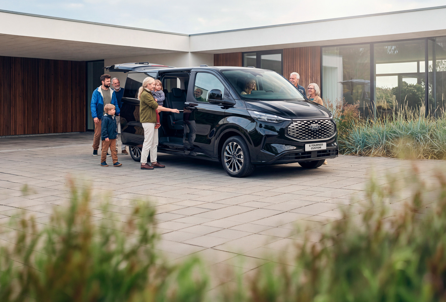 En gruppe mennesker, herunder voksne og børn, står ved siden af en sort Ford E-Tourneo Custom Minibus med sidedøren åben foran et moderne hus.