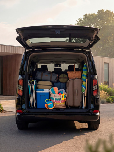 En Ford E-tourneo Custom minivan med bagagerummet åbent, fyldt med bagage og strandudstyr, holder parkeret foran et moderne hus.