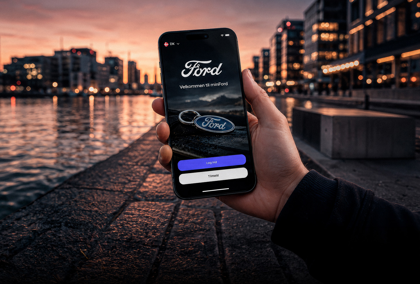 En hånd holder en smartphone, der viser en Ford-loginskærm med et bybillede ved vandet i baggrunden i skumringen.