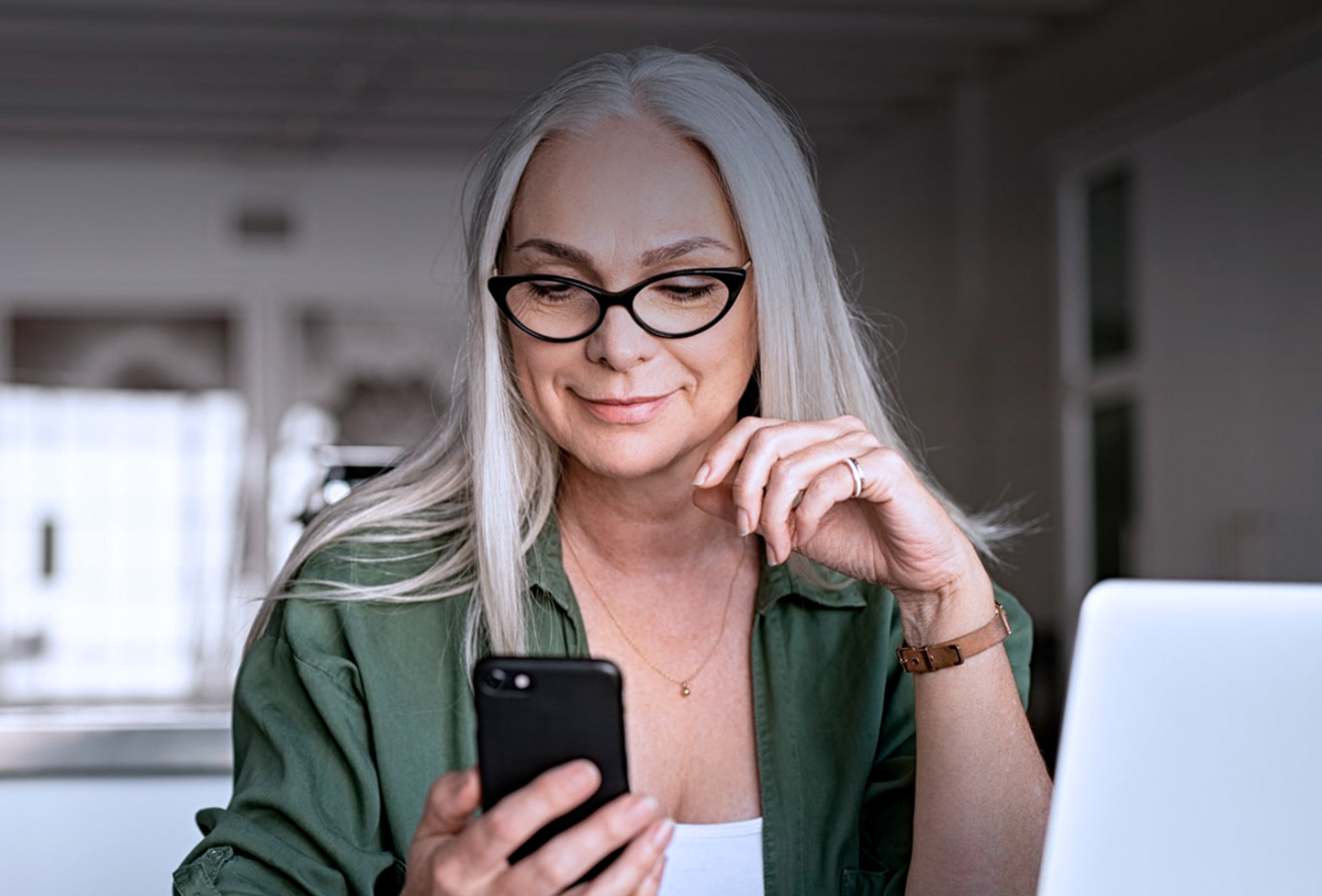 Kvinde med langt gråt hår og briller kigger på sin smartphone, mens hun sidder ved et skrivebord med en bærbar computer.
